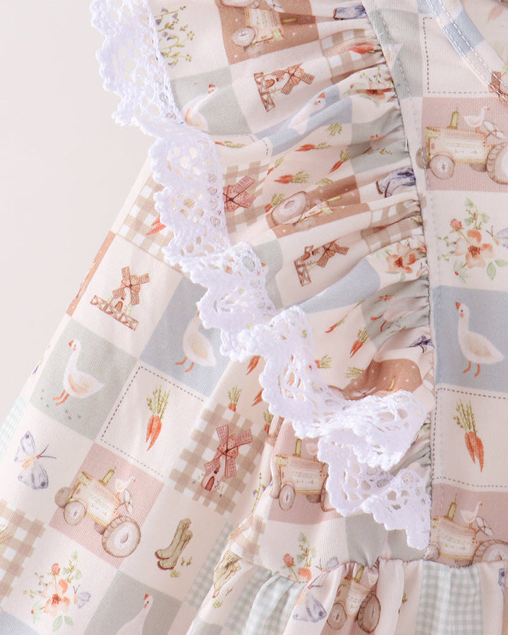 A close-up of a baby dress featuring a patchwork print with white lace ruffles. The print includes illustrations of farm animals, carrots, and tractors in soft pastel colors of pink, blue, and beige.