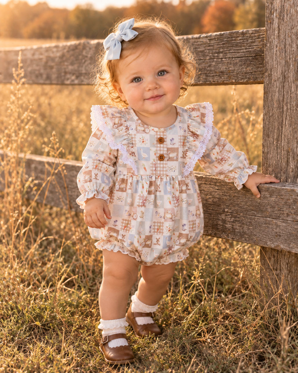 A toddler girl wearing a long-sleeve patchwork romper with ruffled shoulders, lace trim, and three buttons, standing outdoors next to a wooden fence in a grassy field at sunset.