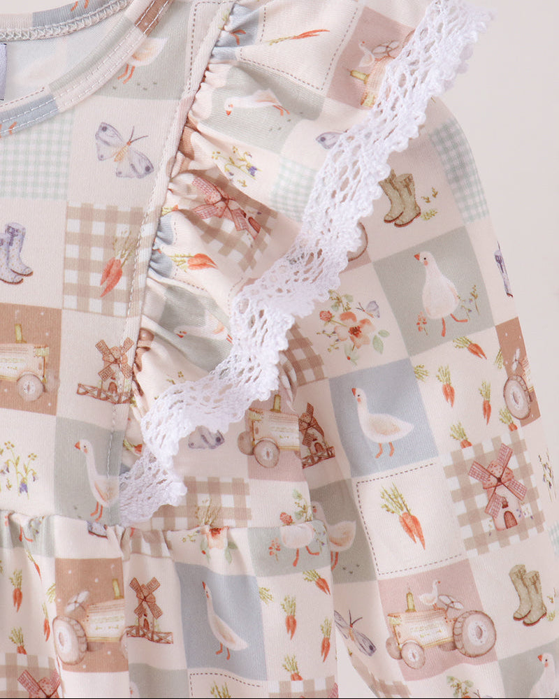 A close-up of a baby dress featuring a patchwork design with cute illustrations of farm animals, carrots, and boots in soft pastel colors, accented by a white lace ruffle.