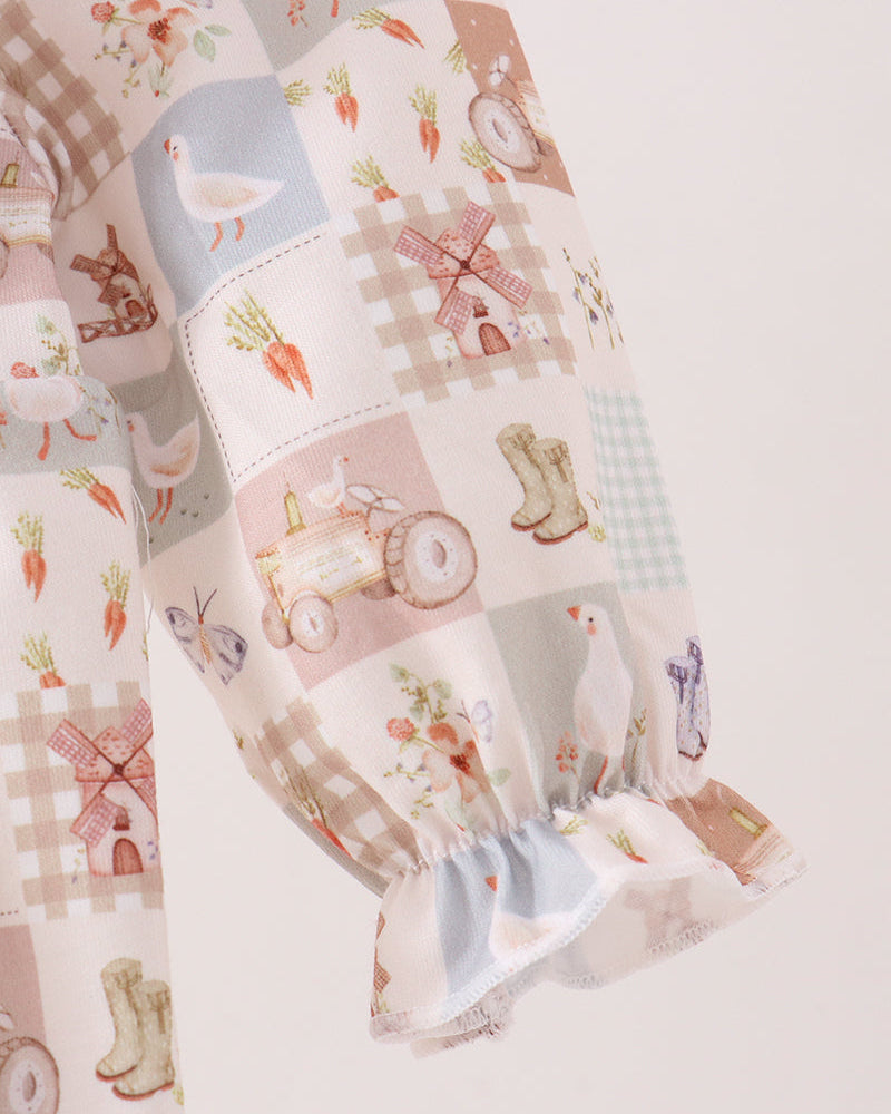 A close-up of a long sleeve featuring a whimsical patchwork print with illustrations of farm animals, carrots, windmills, tractors, and gingham patterns, finished with a ruffled cuff.