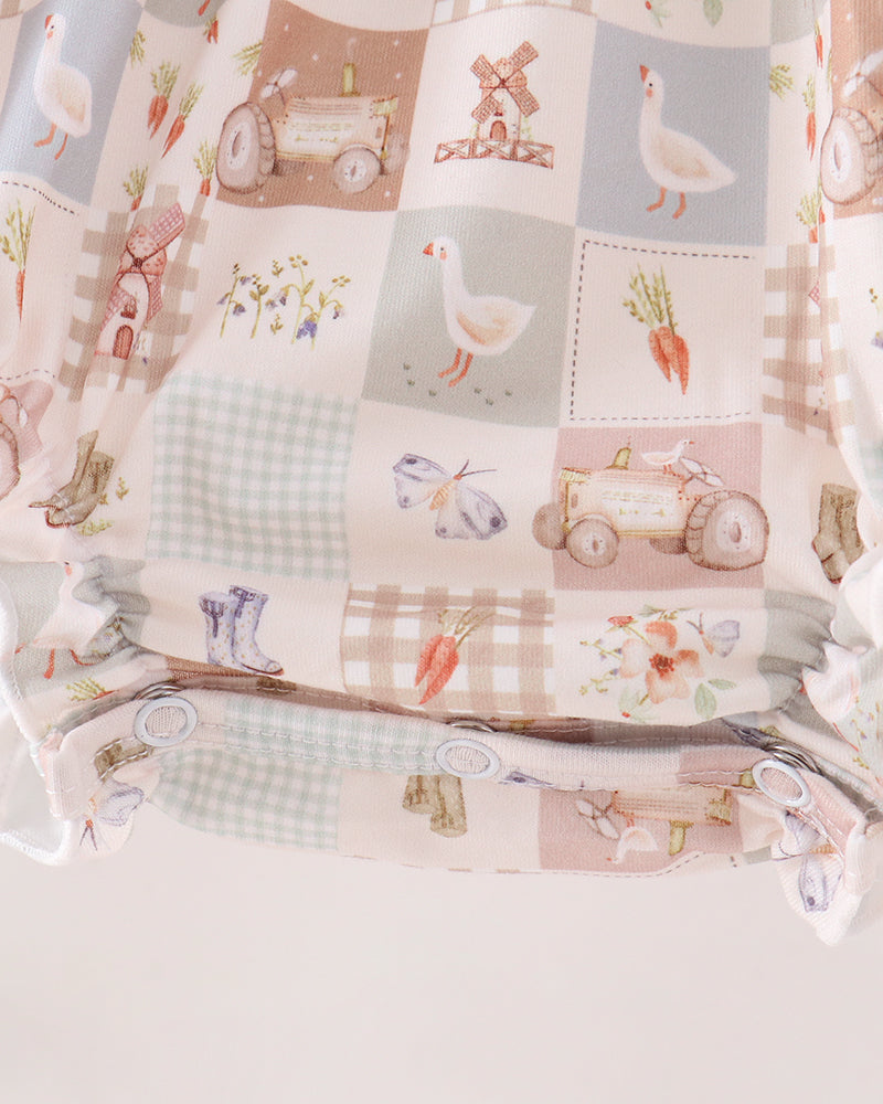 A close-up of a baby diaper or soft fabric accessory with a patchwork-style country farm print. The pattern includes square panels with illustrations of a goose, carrots, a windmill, a tractor, boots, flowers, and gingham checks in muted beige, blush pink, pale blue, and cream tones.