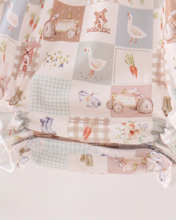 A close-up of a baby diaper or soft fabric accessory with a patchwork-style country farm print. The pattern includes square panels with illustrations of a goose, carrots, a windmill, a tractor, boots, flowers, and gingham checks in muted beige, blush pink, pale blue, and cream tones.