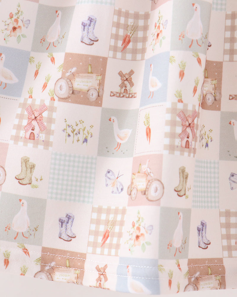 A close-up of a fabric featuring a patchwork design with a rustic farm theme. The pattern includes squares with illustrations of white ducks, purple rain boots, carrots, windmills, tractors, butterflies, and gingham checks in soft pastel colors of pink, blue, beige, and green.