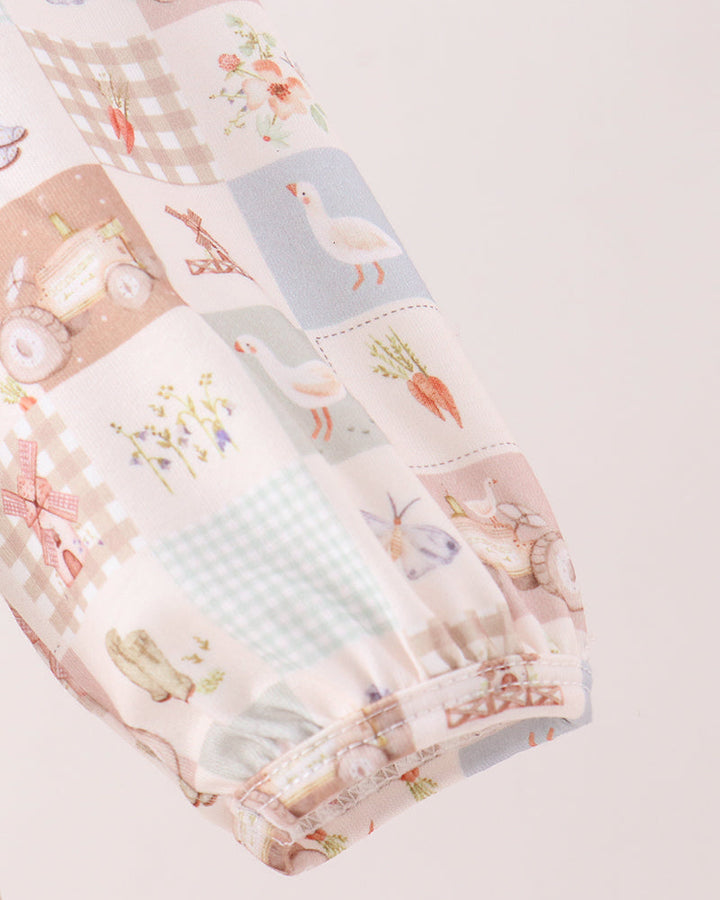 A close-up of a long-sleeve dress featuring a soft pastel patchwork print with gingham, ducks, carrots, and small floral motifs on a pale pink background.