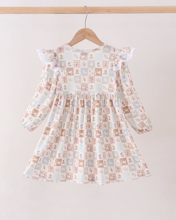 A long-sleeve dress with a ruffled collar and flutter sleeves, featuring a patchwork print of small squares in muted blue, brown, and beige tones. The dress is displayed on a wooden hanger hanging from a wooden rod against a plain off-white wall.