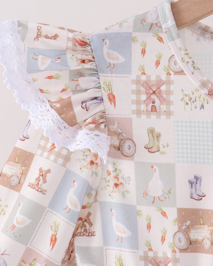 A close-up of a patchwork-style baby dress or top featuring a soft pastel print with ducks, carrots, windmills, rain boots, and floral motifs, plus white lace trim and a wooden hanger.