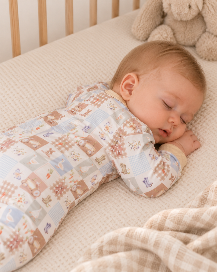 A sleeping baby lying on their side in a crib, wearing a long-sleeve patchwork-patterned sleeper in soft beige, brown, and light blue tones. A beige stuffed bunny and a beige plaid blanket are visible in the scene.