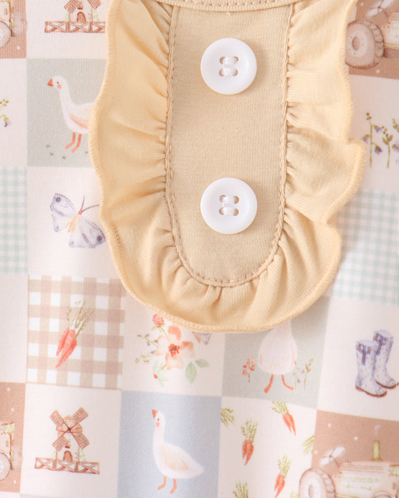 A close-up of a pale yellow baby garment with two white buttons and ruffled edges, laid flat on a patchwork fabric background with farm and animal illustrations.