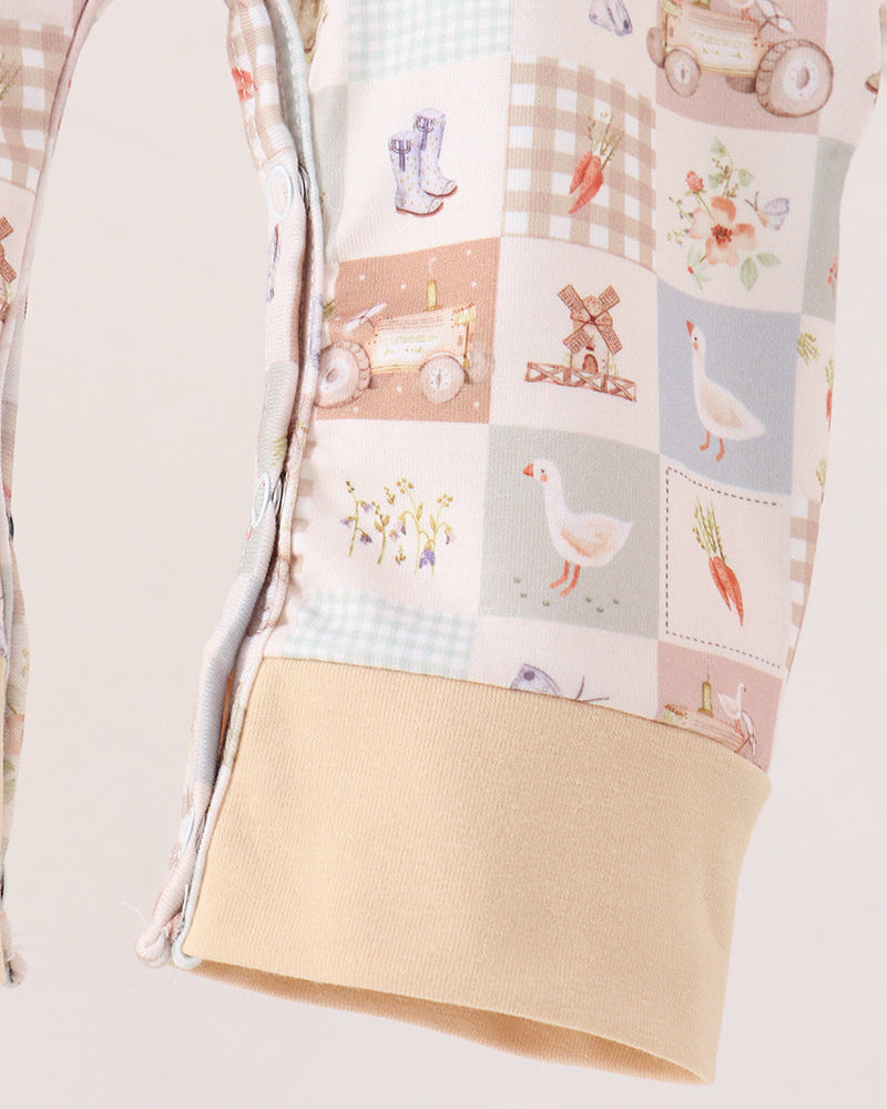 A close-up of a patchwork fabric garment, likely a baby blanket or quilt, featuring a retro farm-themed print with patches of carrots, windmills, geese, and tractors in soft pastel colors, finished with a solid beige cuff.