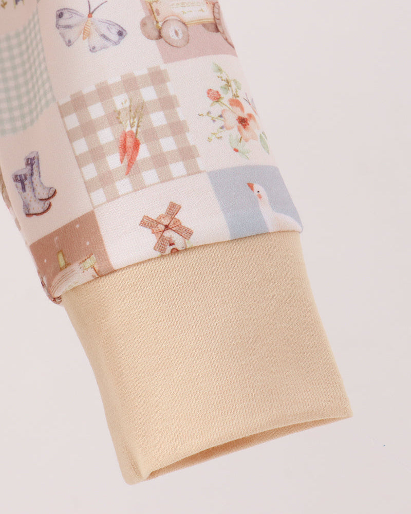 Close-up of a sleeve cuff featuring a patchwork print with pastel illustrations of carrots, a windmill, flowers, gingham, and checkered patterns, finished with a solid beige ribbed knit cuff.