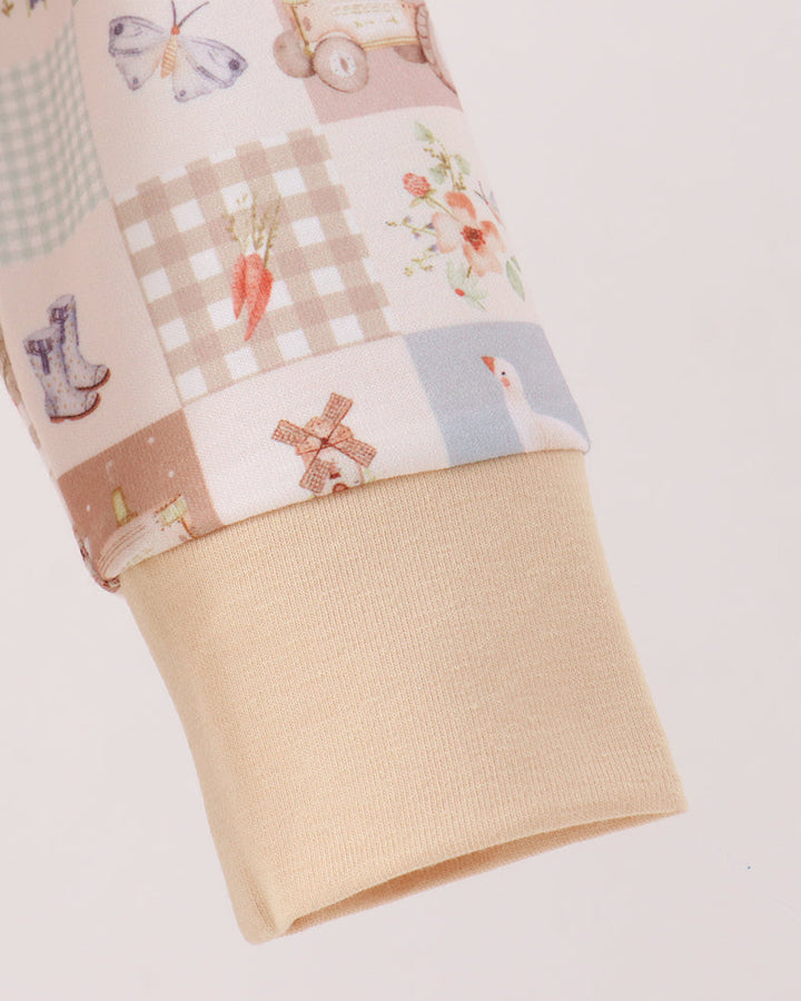 Close-up of a sleeve cuff featuring a patchwork print with pastel illustrations of carrots, a windmill, flowers, gingham, and checkered patterns, finished with a solid beige ribbed knit cuff.