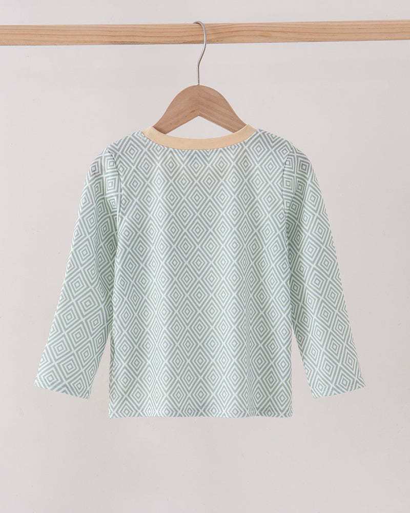 A long-sleeve boys top with a light mint green and white geometric diamond pattern, hanging on a wooden hanger from a wooden clothes rack against a plain light beige background.