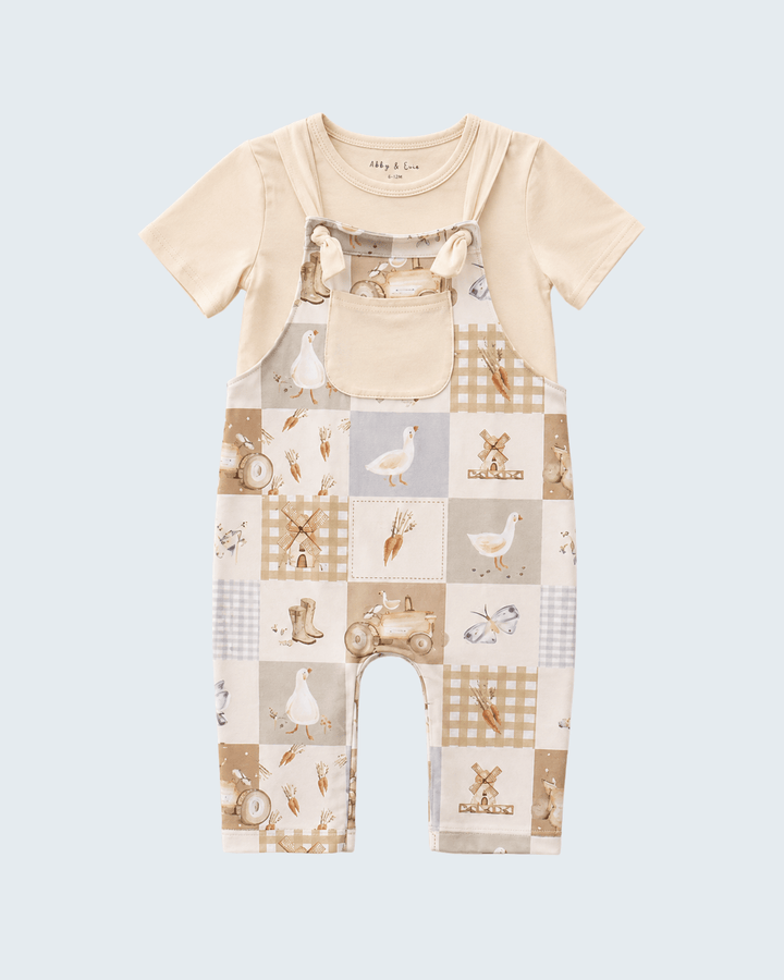 A baby short-sleeve romper in a cream and beige patchwork print featuring gingham, stripes, and illustrations of farm animals and vehicles, shown against a light blue-gray background.