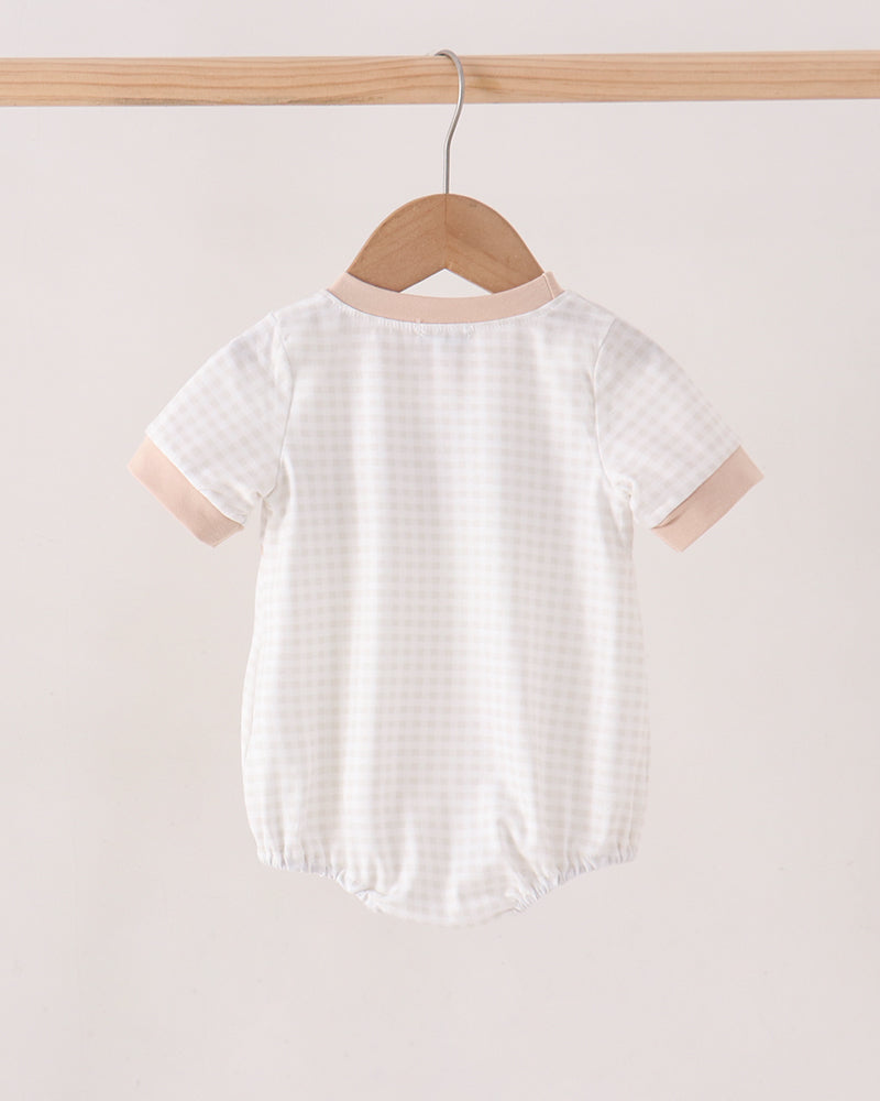 A short-sleeve baby romper hanging on a wooden hanger from a light wood rod, featuring a white and beige gingham pattern with solid beige trim on the collar and sleeve cuffs.