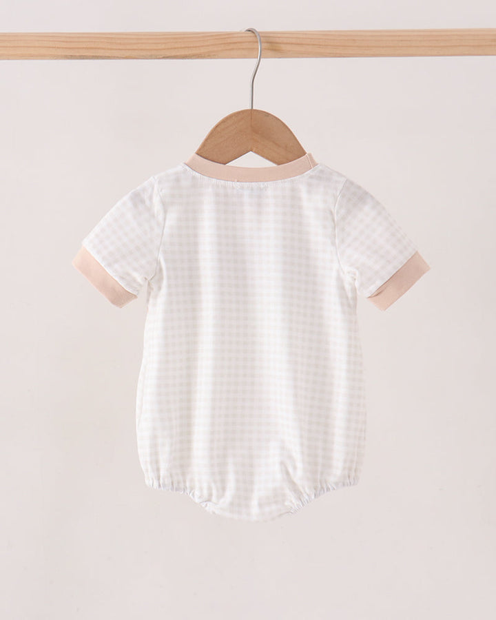 A short-sleeve baby romper hanging on a wooden hanger from a light wood rod, featuring a white and beige gingham pattern with solid beige trim on the collar and sleeve cuffs.