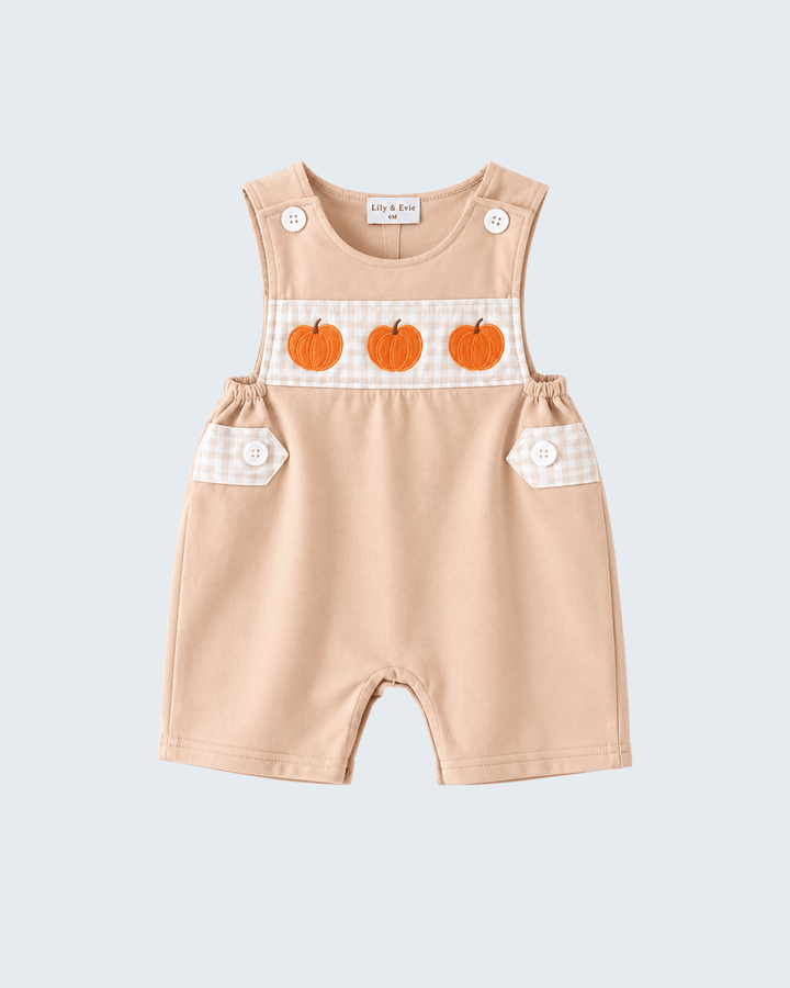 A baby romper in a light peach-beige color, featuring three orange embroidered pumpkins on a white gingham patterned chest panel. The romper has buttoned pockets with matching gingham fabric and white snap buttons on the shoulder straps, shown against a solid light gray background.