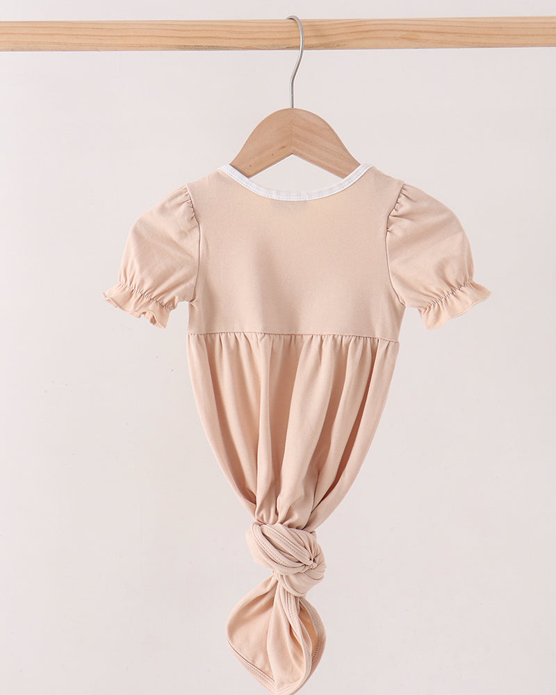A light beige short-sleeve baby dress with a gathered waist and knotted bottom, hanging on a wooden hanger from a wooden clothes rack against a plain off-white background.