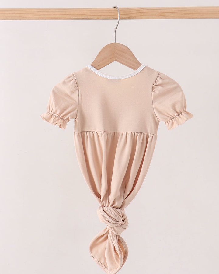 A light beige short-sleeve baby dress with a gathered waist and knotted bottom, hanging on a wooden hanger from a wooden clothes rack against a plain off-white background.
