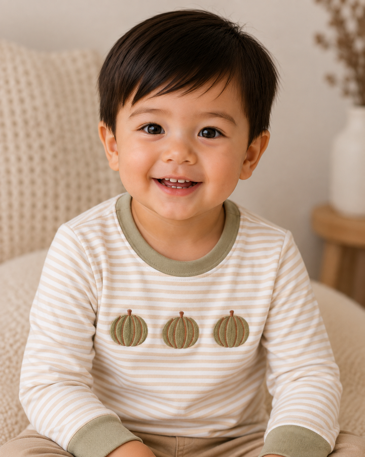 A smiling toddler boy wearing a cream and beige horizontally striped long-sleeve shirt with three olive green embroidered pumpkins on the chest, sitting on a cozy beige knitted chair.