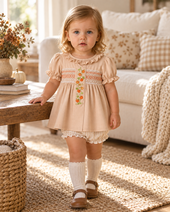 A toddler girl wearing a beige short-sleeve smocked dress with floral embroidery, white ribbed socks, and brown Mary Jane shoes, standing in a cozy, sunlit living room.