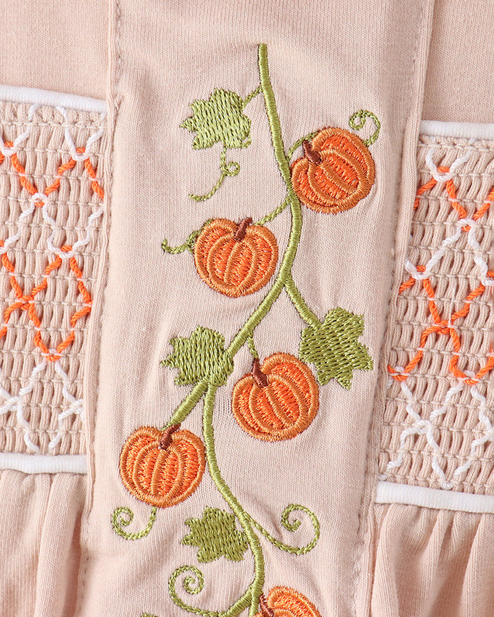Close-up of a light blush pink garment featuring embroidered orange pumpkins on a green vine with green leaves, flanked by white and orange smocked lace panels.