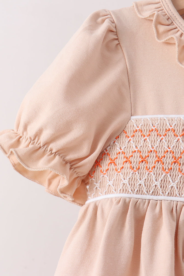 Close-up of a baby dress in a soft peach-beige color, featuring ruffled short puff sleeves and a smocked bodice with orange and white stitching.