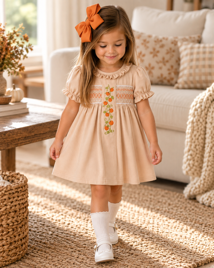 A young girl wearing a beige short-sleeve dress with orange and white floral embroidery on the chest, walking on a woven rug in a cozy living room setting. She is wearing a large orange bow in her hair, white socks, and white shoes.
