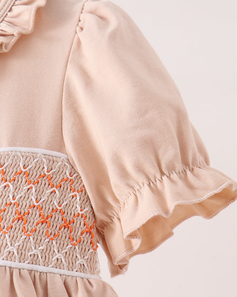Close-up of a light peach dress with ruffled short sleeves, a smocked waist panel in beige and orange, and a gathered skirt, shown against a plain light background.