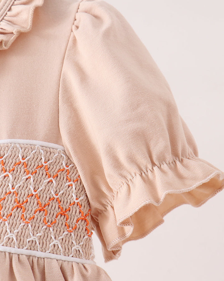 Close-up of a light peach dress with ruffled short sleeves, a smocked waist panel in beige and orange, and a gathered skirt, shown against a plain light background.