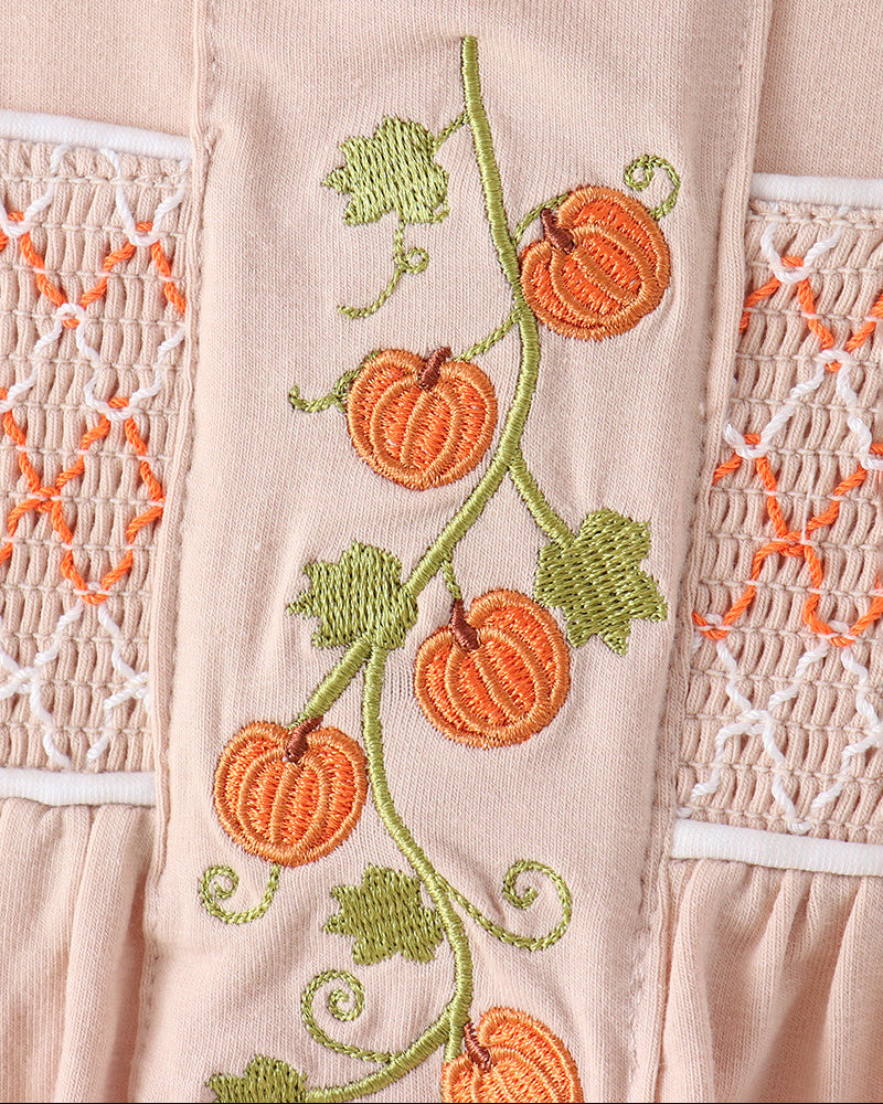Close-up of a light blush pink garment featuring embroidered orange pumpkins on a green vine with green leaves, flanked by white and orange smocked lace panels.