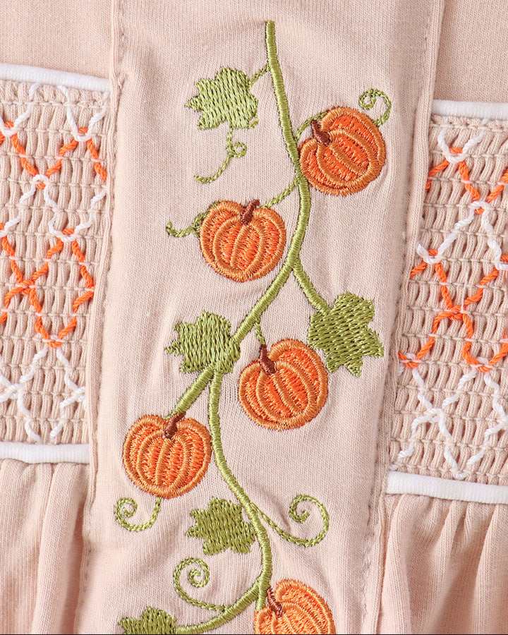 Close-up of a light blush pink garment featuring embroidered orange pumpkins on a green vine with green leaves, flanked by white and orange smocked lace panels.