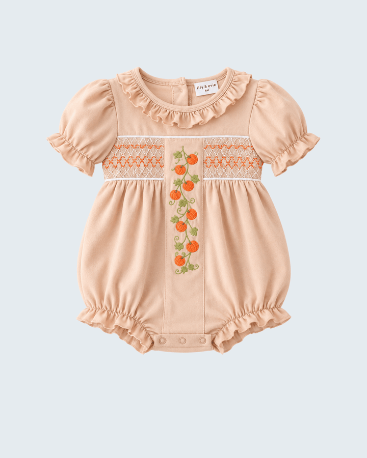 A baby romper in a soft peach-beige color, featuring short puff sleeves, a ruffled collar, and a smocked chest panel with orange and green citrus embroidery. The brand label reads &