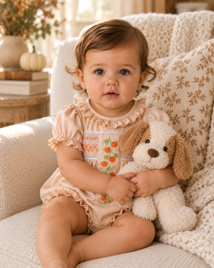 A baby girl sitting on a cozy cream-colored armchair, wearing a peach short-sleeve romper with a floral embroidered chest panel and holding a plush cream and brown dog toy.