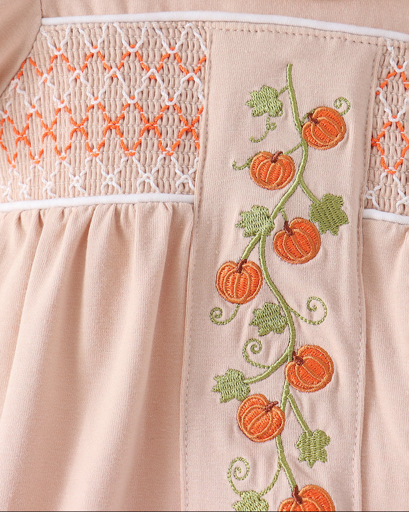 Close-up of a light peach dress featuring orange pumpkin embroidery on a green vine with leaves, alongside white smocked lace detailing.