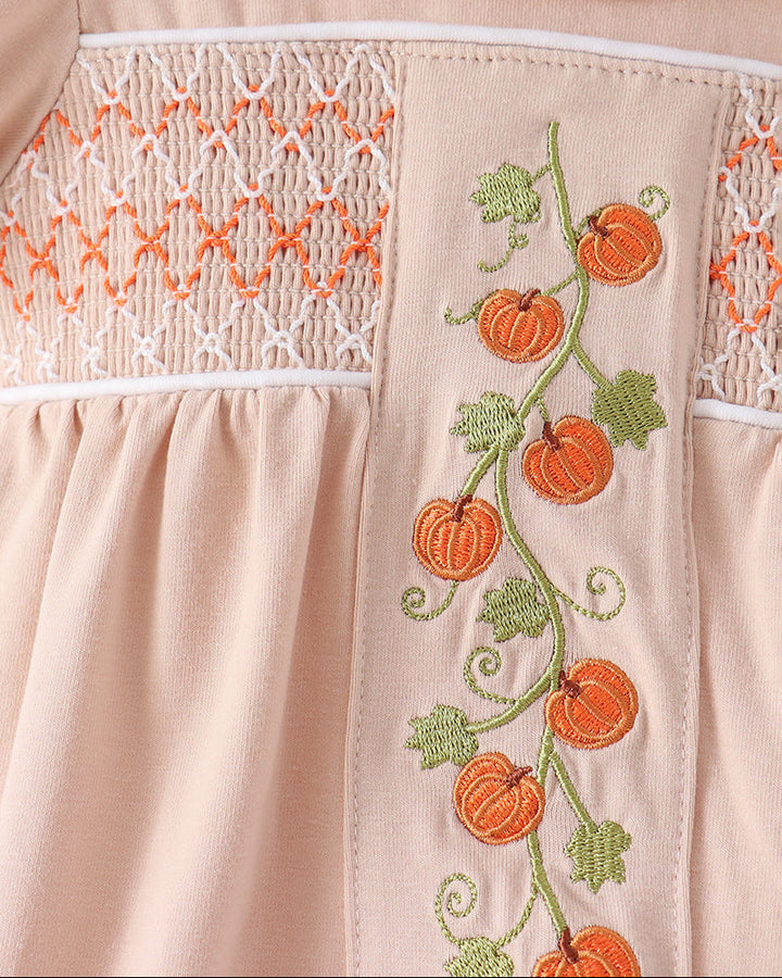 Close-up of a light peach dress featuring orange pumpkin embroidery on a green vine with leaves, alongside white smocked lace detailing.