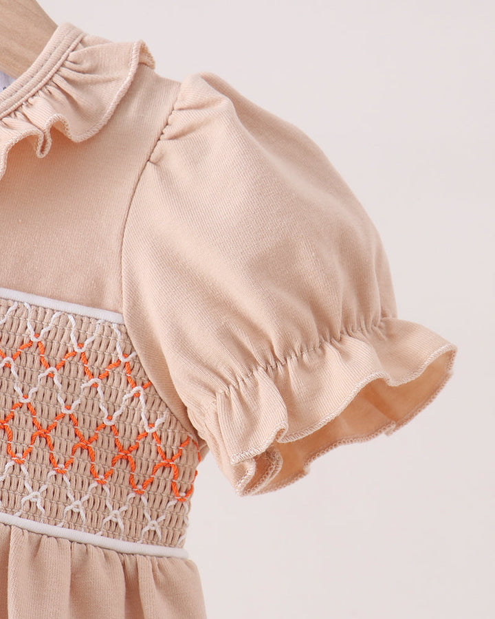 Close-up of a baby dress in a light peach-beige color, featuring a smocked bodice with orange and white embroidery and ruffled short sleeves.
