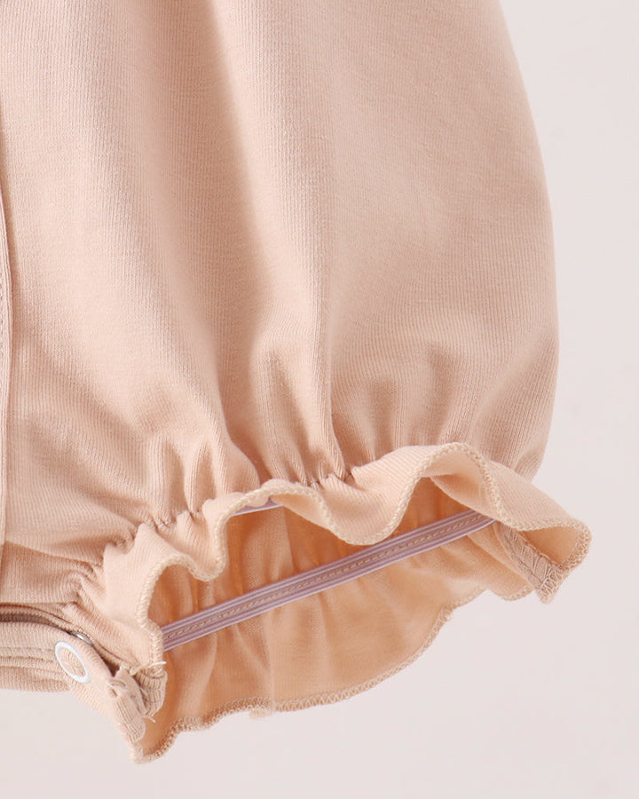 Close-up of a light peach-beige fabric garment with a gathered elastic ruffle hem and a small white snap detail, shown against a plain light background.