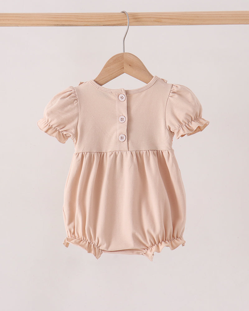 A baby romper in a soft, pale peach-beige color, hanging on a wooden hanger from a wooden clothes rack. The romper features short puff sleeves, a buttoned neckline with three buttons, and a gathered skirt with ruffled trim at the legs.