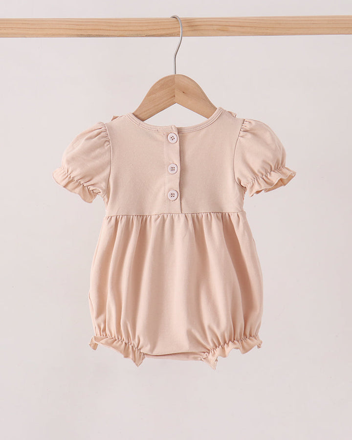 A baby romper in a soft, pale peach-beige color, hanging on a wooden hanger from a wooden clothes rack. The romper features short puff sleeves, a buttoned neckline with three buttons, and a gathered skirt with ruffled trim at the legs.