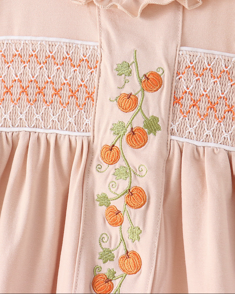 Close-up of a light peach baby dress featuring orange pumpkin and green leaf embroidery down the center, flanked by white smocked lace panels.