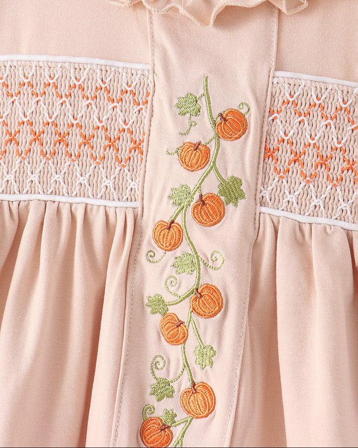 Close-up of a light peach baby dress featuring orange pumpkin and green leaf embroidery down the center, flanked by white smocked lace panels.