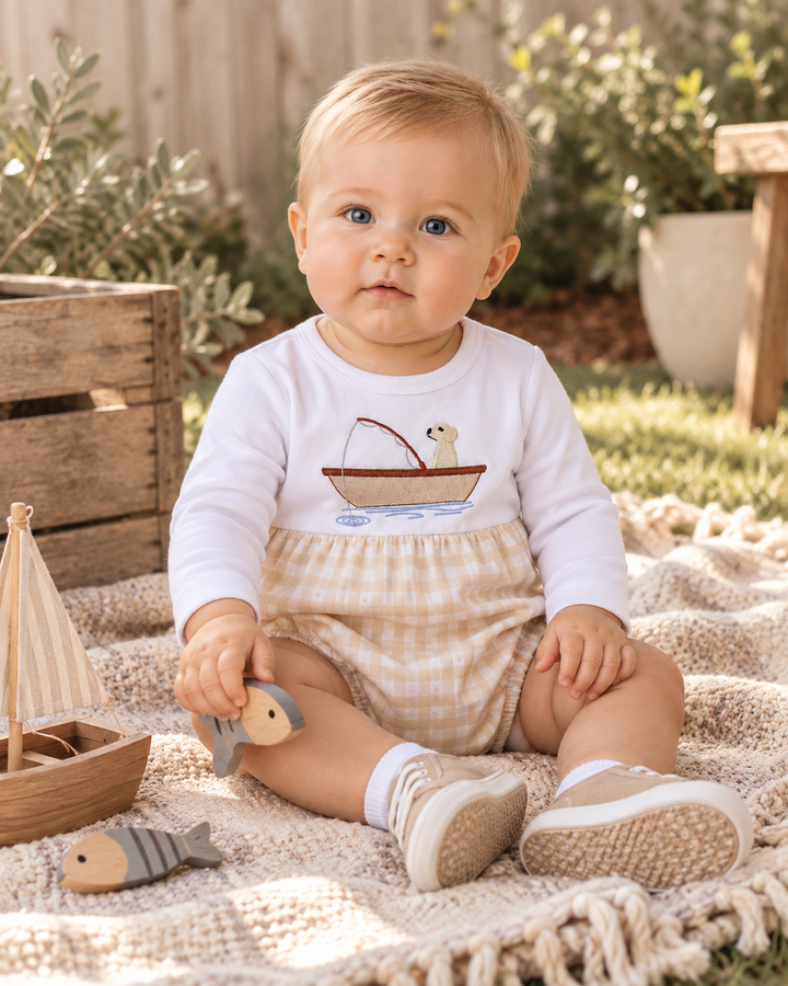 A baby sitting outdoors on a knitted blanket, wearing a white long-sleeve shirt with a fishing boat graphic and beige gingham shorts.