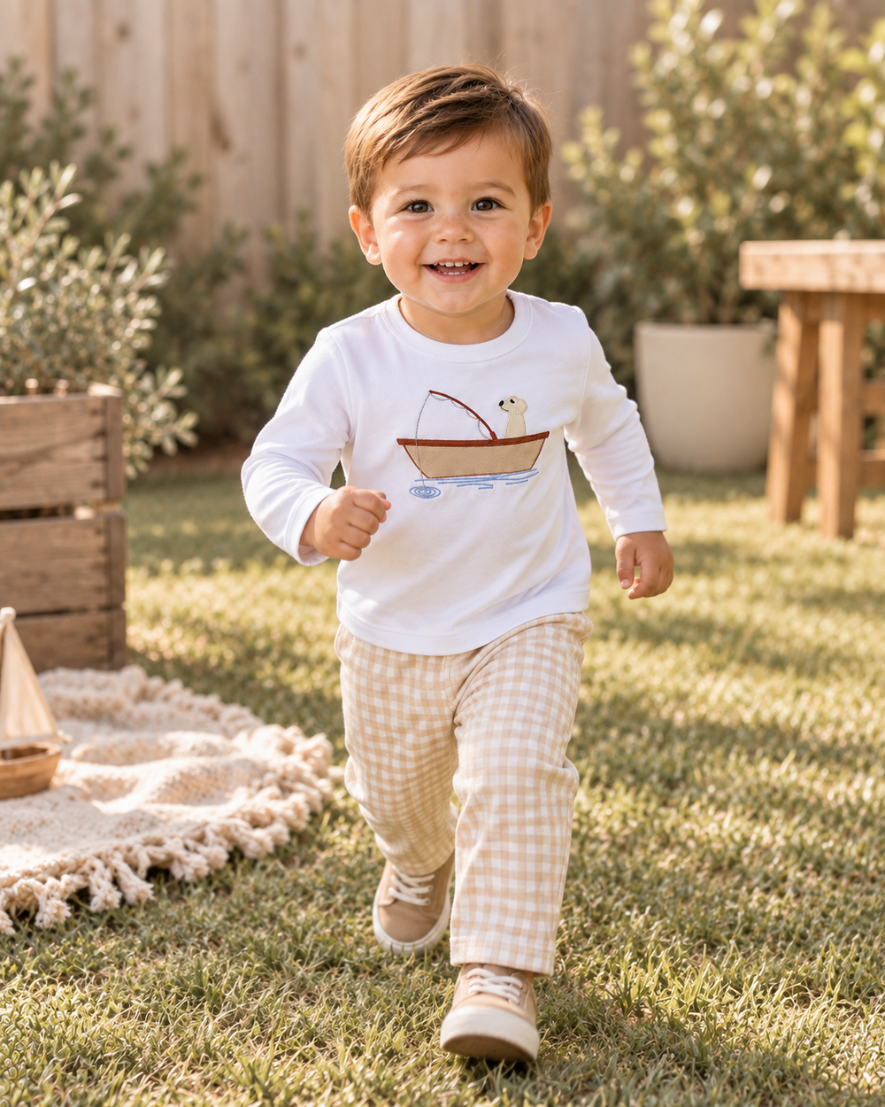 A toddler boy smiling and running outdoors on a grassy lawn, wearing a white long-sleeve shirt with a small boat graphic and beige-and-white gingham pants.