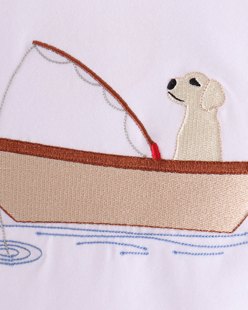 Embroidered design of a cream-colored dog sitting in a beige and brown boat on a white fabric background.