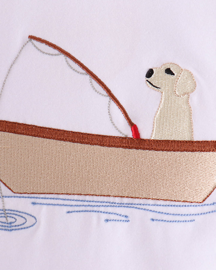Embroidered design of a cream-colored dog sitting in a beige and brown boat on a white fabric background.
