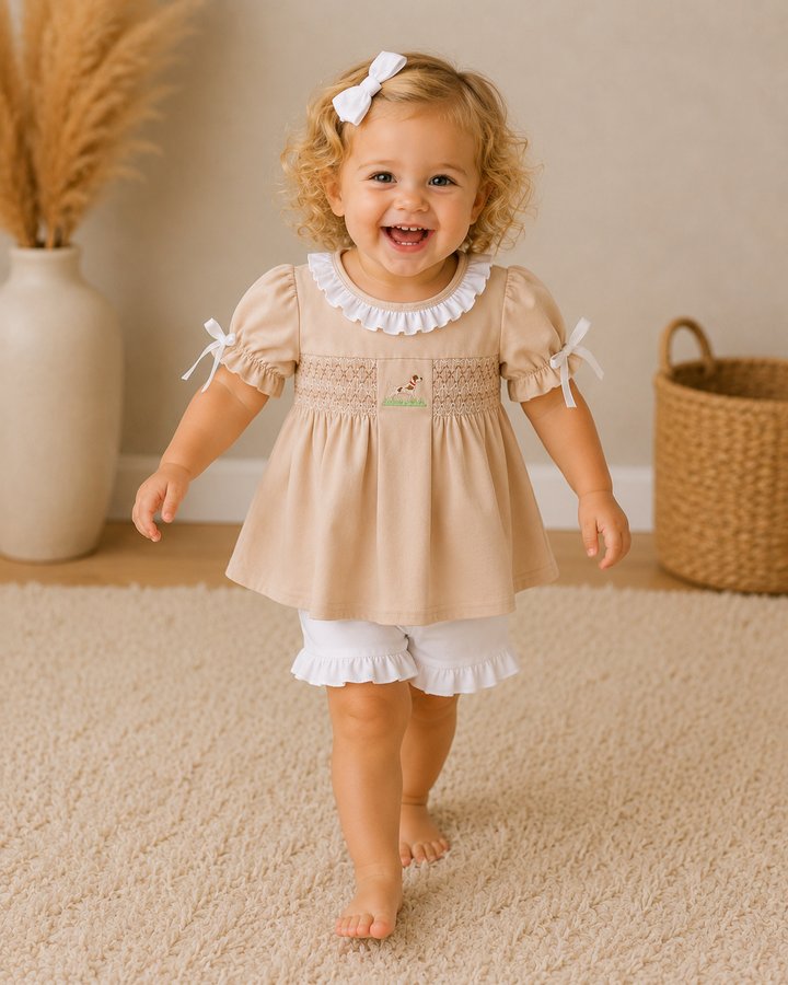 Child wearing a beige dress with white ruffled collar and hem, standing on a carpeted floor.