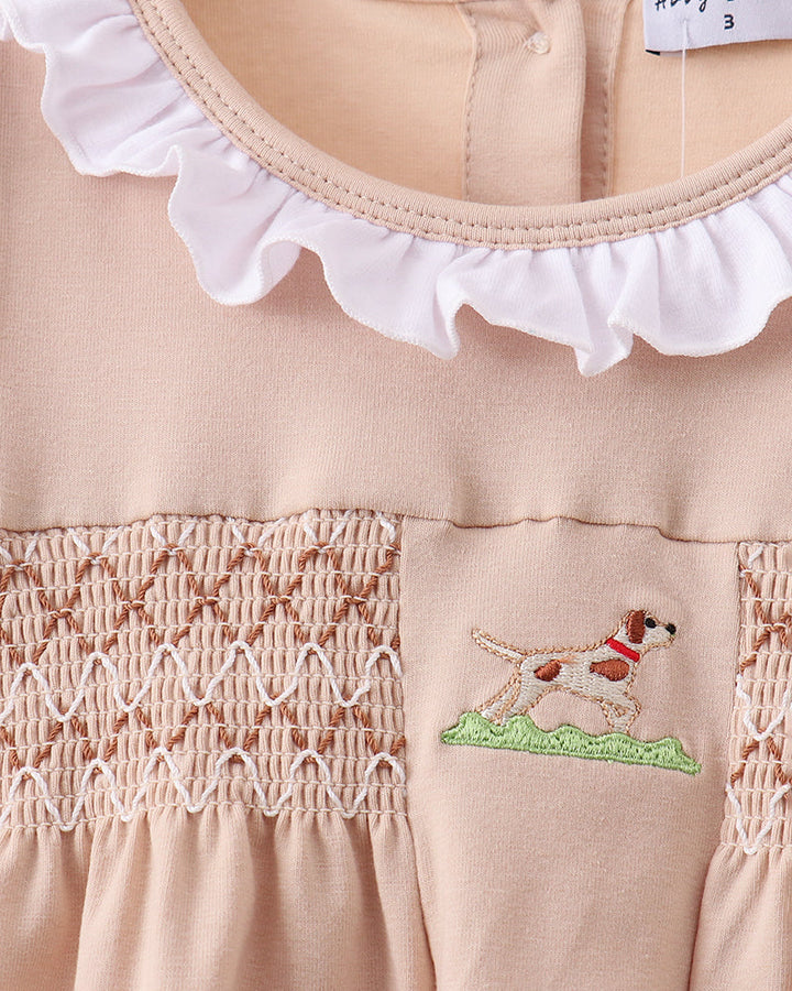 Close-up of a pink dress with smocked details and a dog embroidery on a beige background.