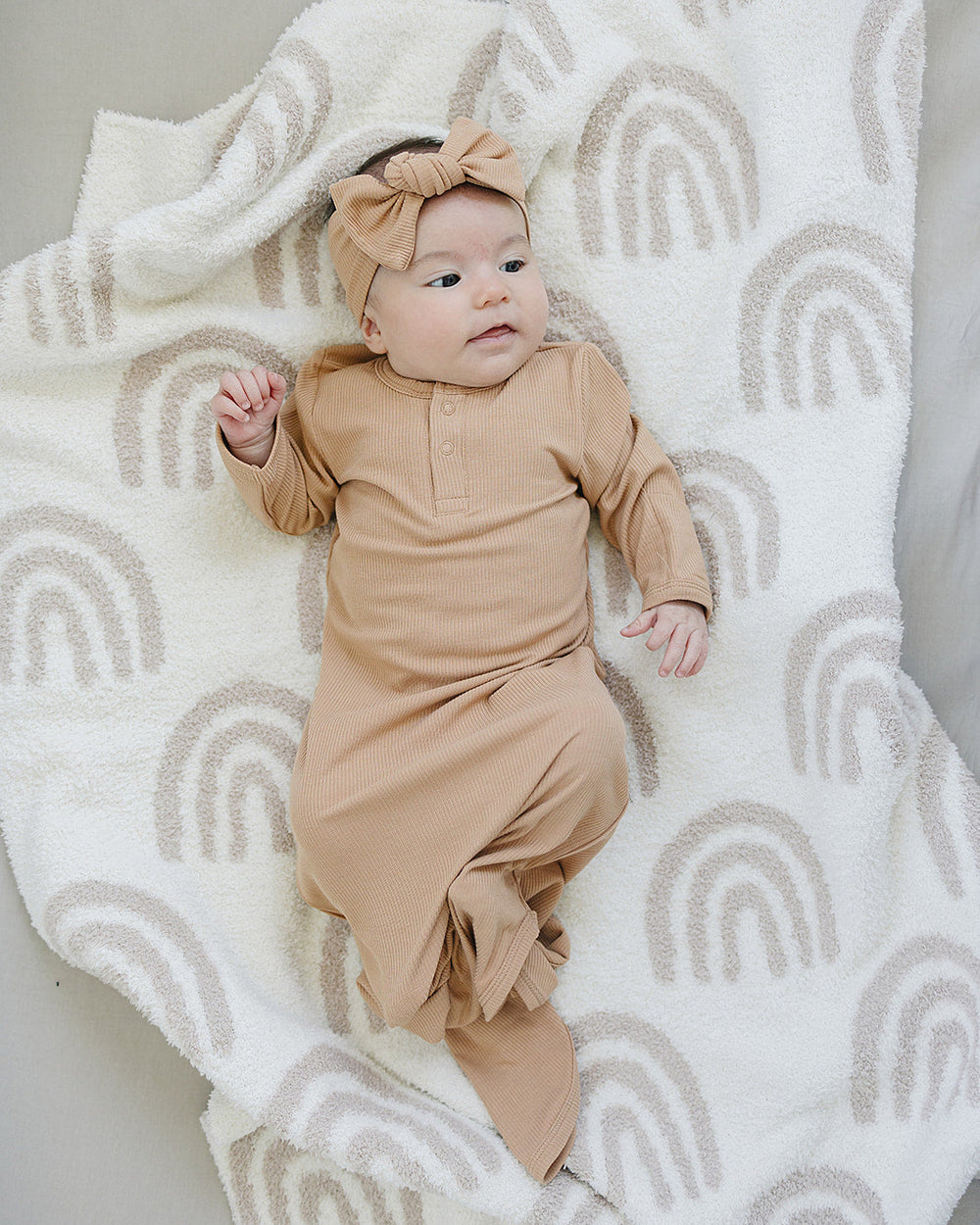 Baby in a beige outfit lying on a white blanket with rainbow patterns