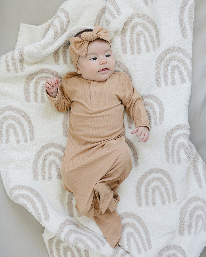 Baby in a beige outfit lying on a white blanket with rainbow patterns