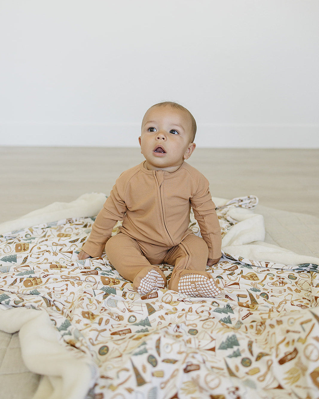 Baby in a brown onesie sitting on a patterned blanket against a white wall